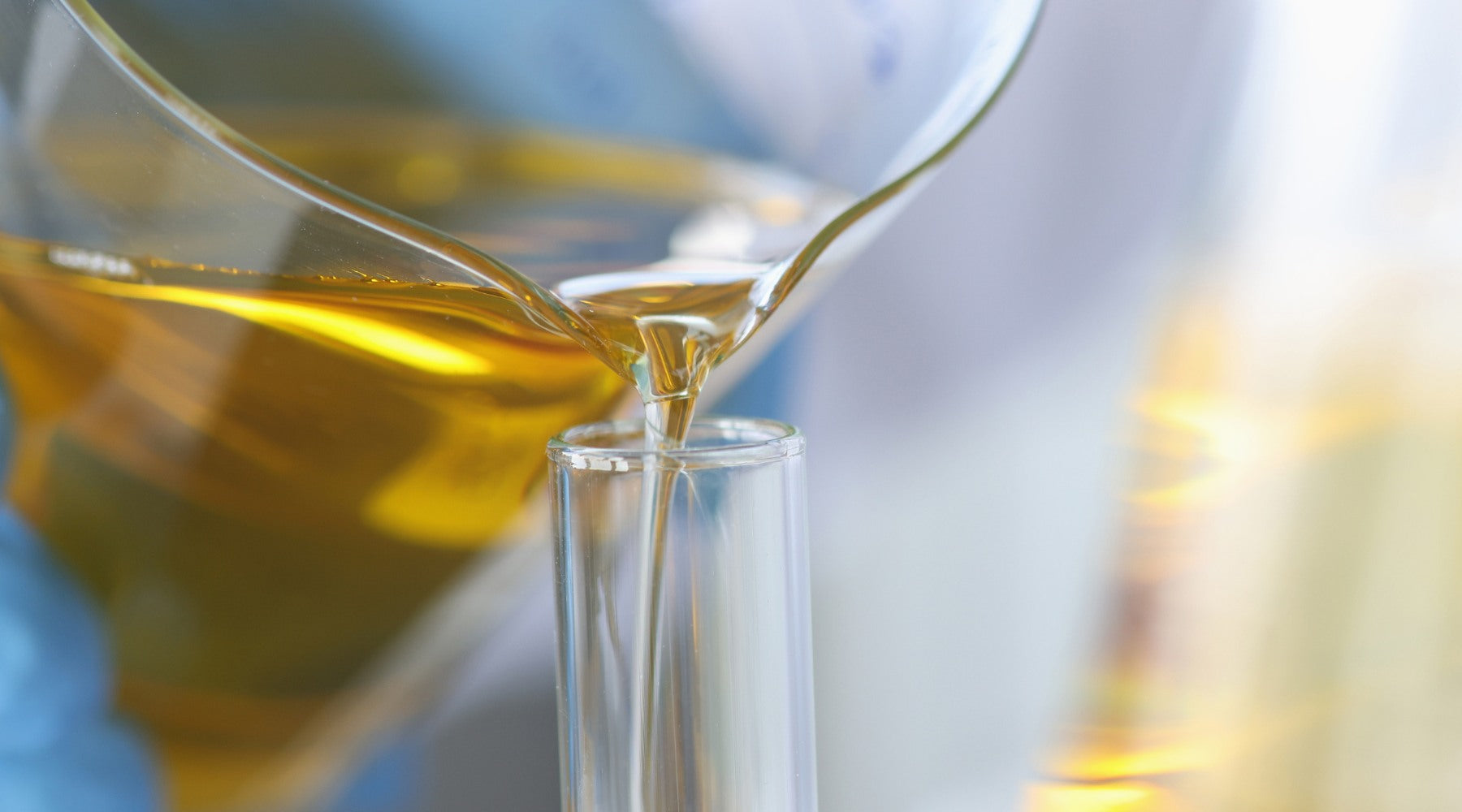 A slightly blurry view of a hand wearing blue laboratory gloves pouring golden jojoba oil from a beaker into a glass tube. The beaker is positioned in the upper left corner, with the glass tube in the center.