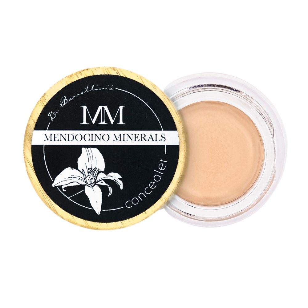 Dr. Berrettini's Mendocino Minerals 'Nude Beach' concealer, a light to light-medium shade, in a top-down view. Shows glass bottom packaging and a bamboo lid with a black label, placed partially over the container but off to the side.